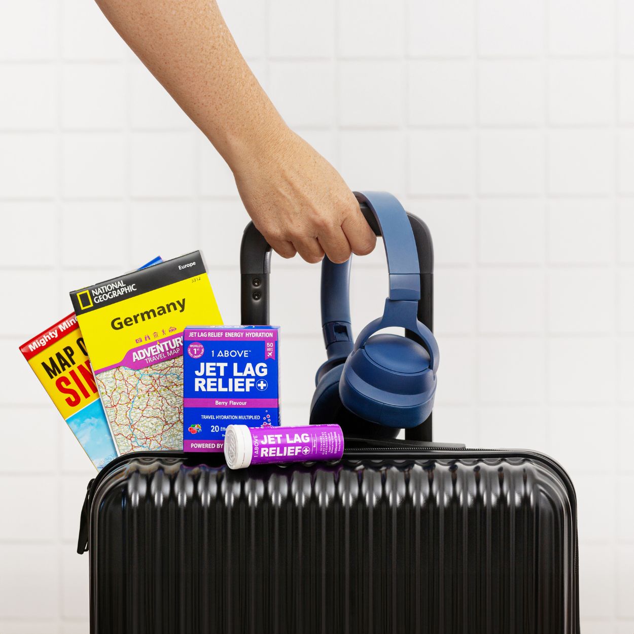 Black suitcase with travel items including headphones, maps, and a 1Above Jet Lag Relief on a white background.